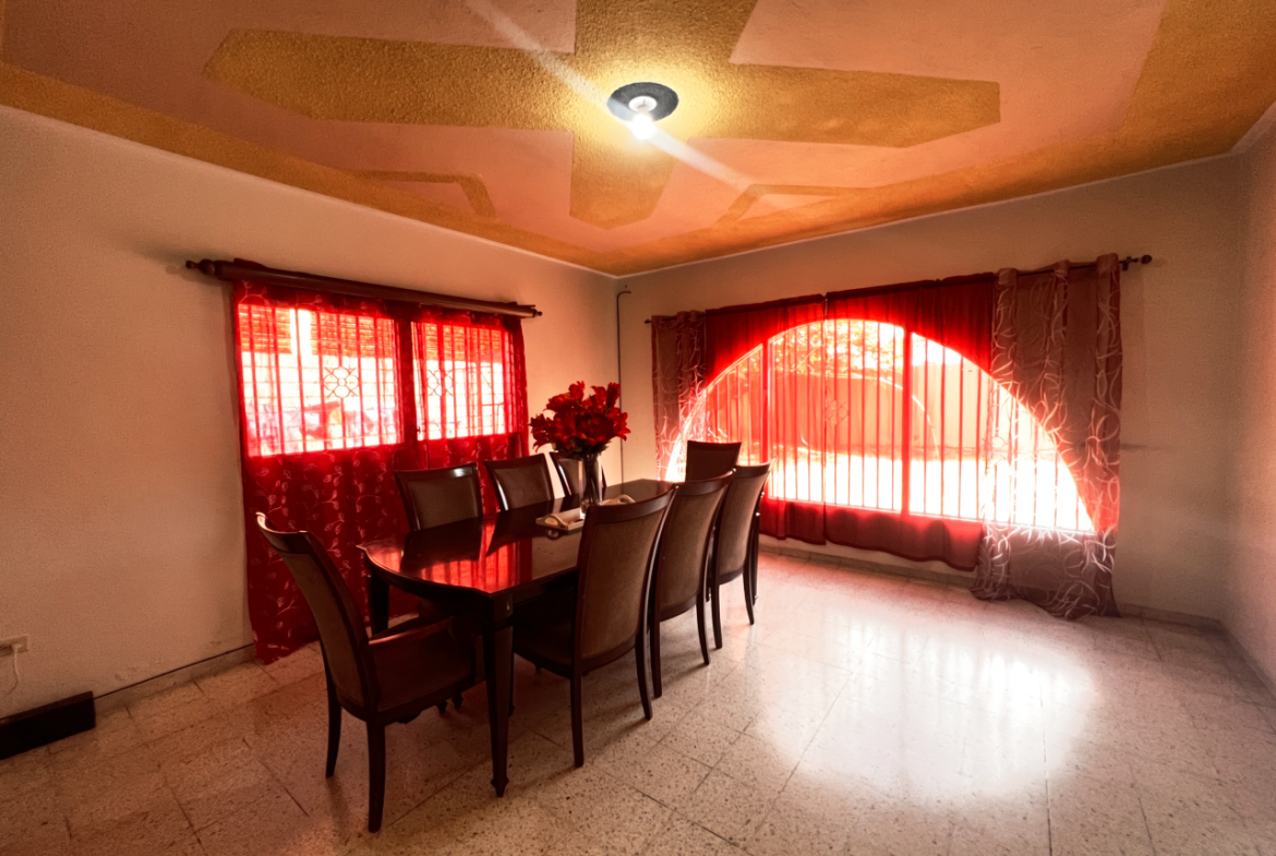 Sala de comedor familiar, contiene 2 ventanas con cortinas color rojo con vista al patio de la casa.