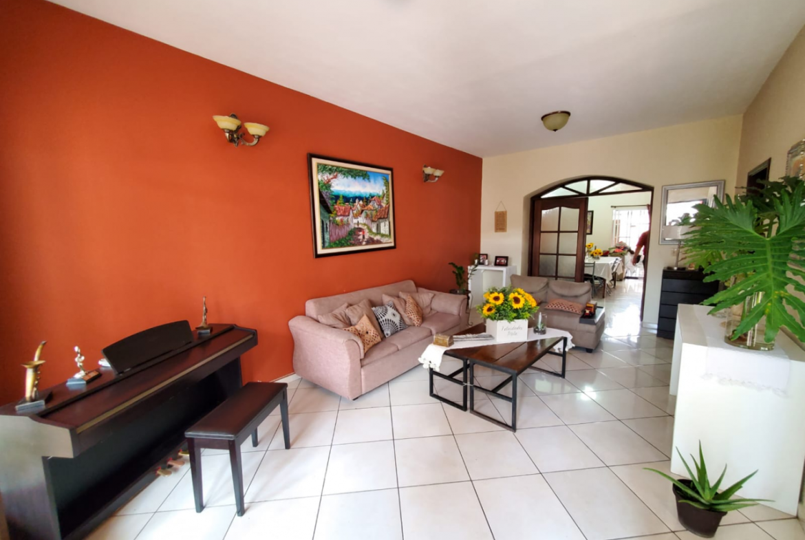 Sala familiar que contiene dos sofás, una mesa de centro con un arreglo de flores encima, un piano, la puerta principal con acceso al jardín en Tegucigalpa, Honduras.