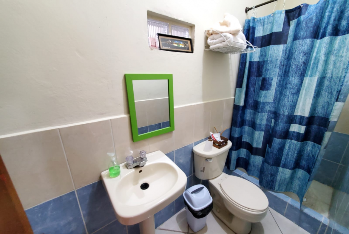 Baño con su inodoro, lavamanos, espejo, una estante para poner toallas y una cortina de baño azul en la colonia Modelo, Honduras.