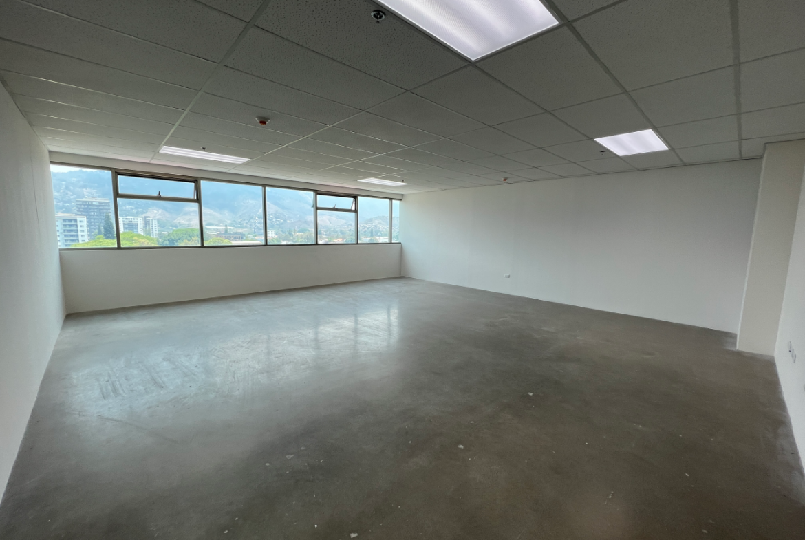 Espacio de oficina con piso de cemento sólido, paredes y techo de tabla yeso color blanco, lámparas led rectangulares empotradas y una ventana larga horizontal con vista a la ciudad.