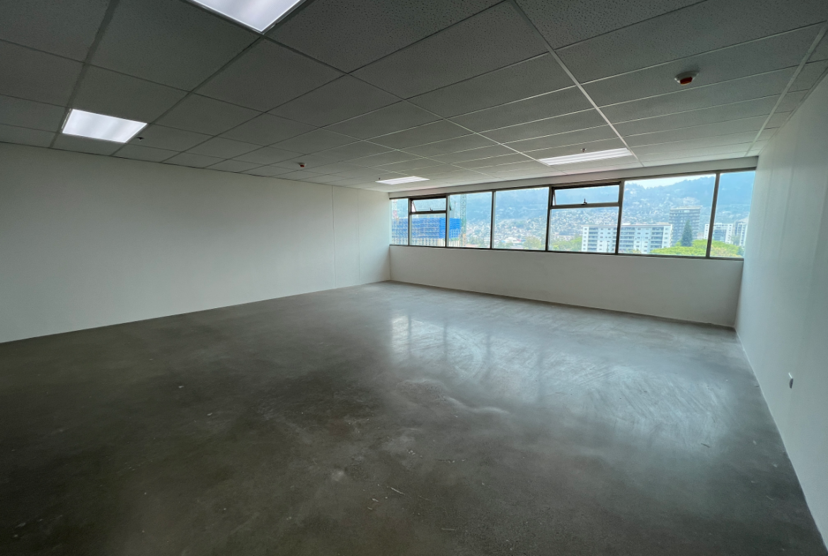 Espacio de oficina en torre agalta con piso de cemento sólido, paredes y techo tabla yeso color blanco, lámparas led rectangulares empotradas y una ventana larga horizontal con vista a la ciudad.