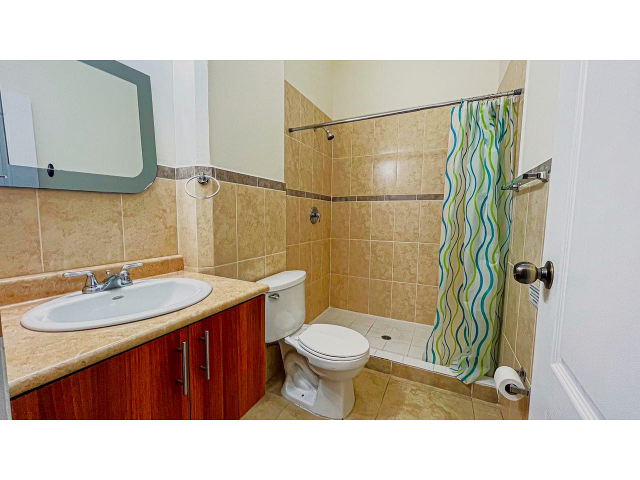 Baño con paredes de color blanco, suelo de cerámica, regadera de cerámica con cortina de protección, baño y lavamanos de color blanco, con pequeño espejo.