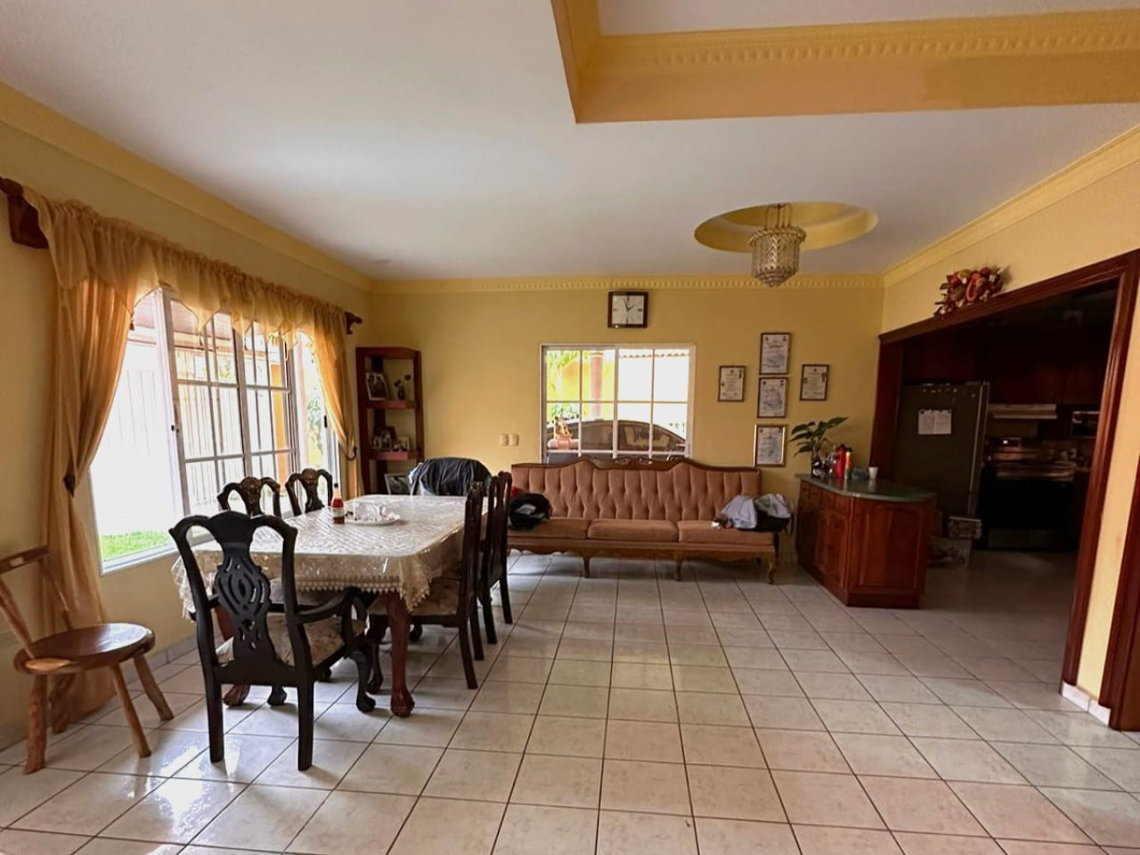 Area de comedor con con paredes de color amarillo y suelo de ceramica blanca, mesa de comedor de madera con 6 sillas de madera oscura, ventanas que dan vista al jardín.
