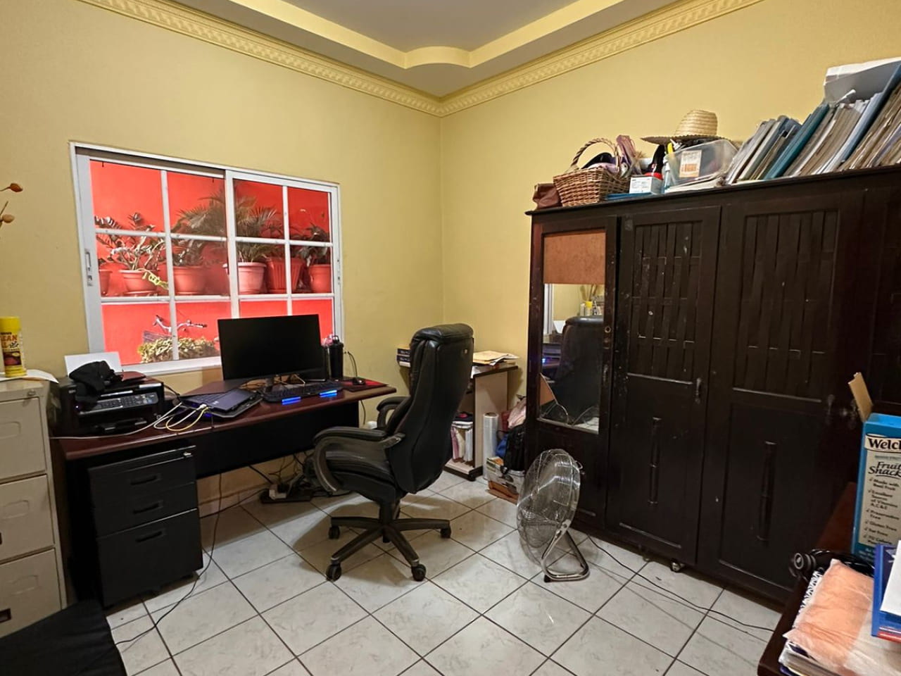 Área de oficina con paredes de color amarillo, con suelo de ceramica color blanco, armario de madera colorcafe oscuro, mesa de oficina con silla a juego.