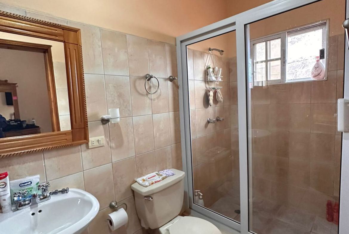 Área de baño con regadera protegida por puera de vidrio deslizable, servicio y lavamanos de color blanco a juego.