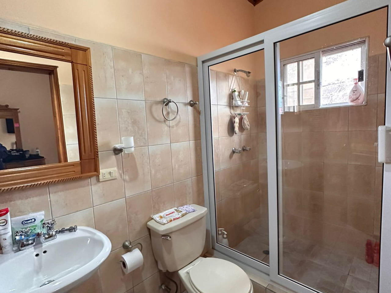 Área de baño con regadera protegida por puera de vidrio deslizable, servicio y lavamanos de color blanco a juego.