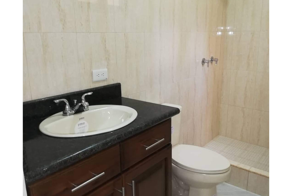 Baño con mueble color café oscuro, plancha color negro en lavamanos junto a inodoro con cerámica en la pared de color blanco.