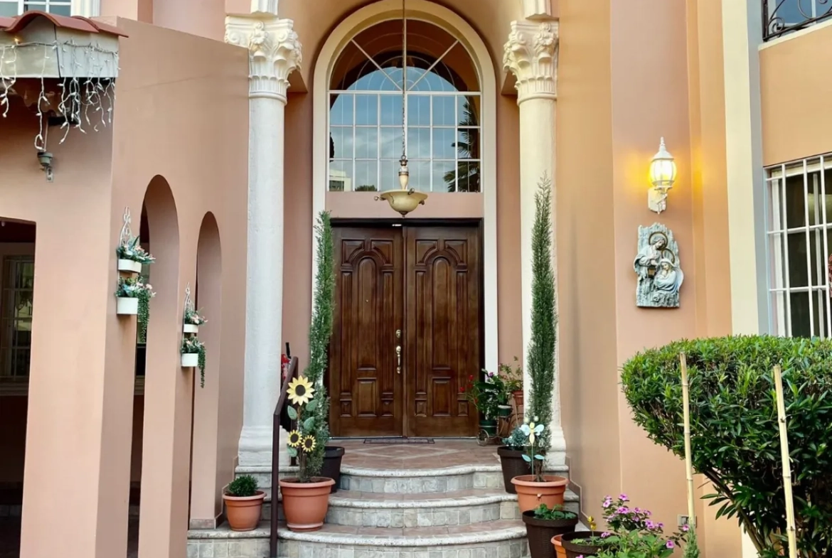 puerta principal doble, hecha de madera con 3 escalones, dos columnas a cada lado, parede de color rosa palido, decorado con diferentes plantas.