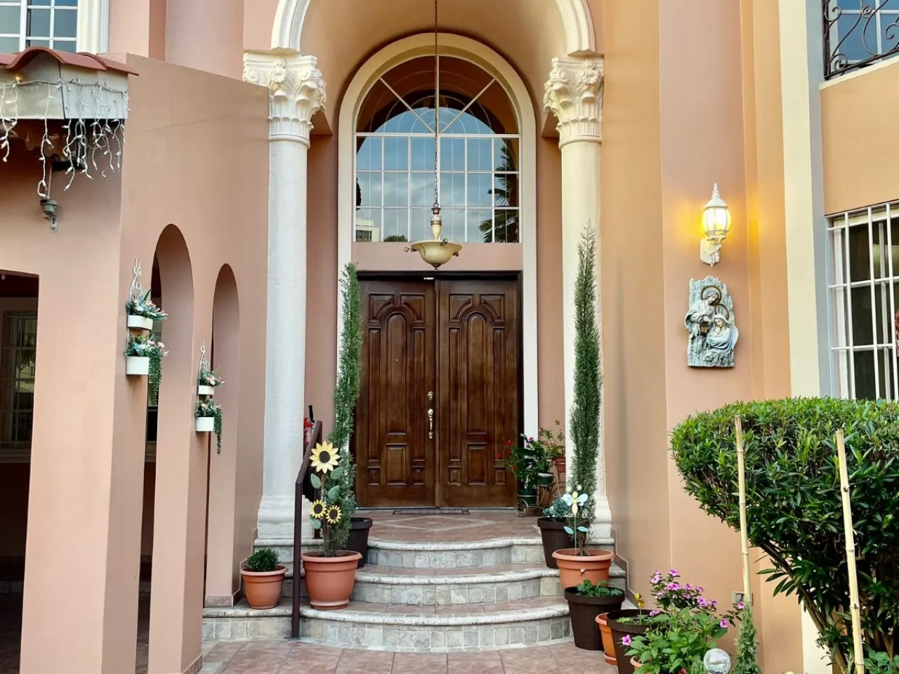 puerta principal doble, hecha de madera con 3 escalones, dos columnas a cada lado, parede de color rosa palido, decorado con diferentes plantas.