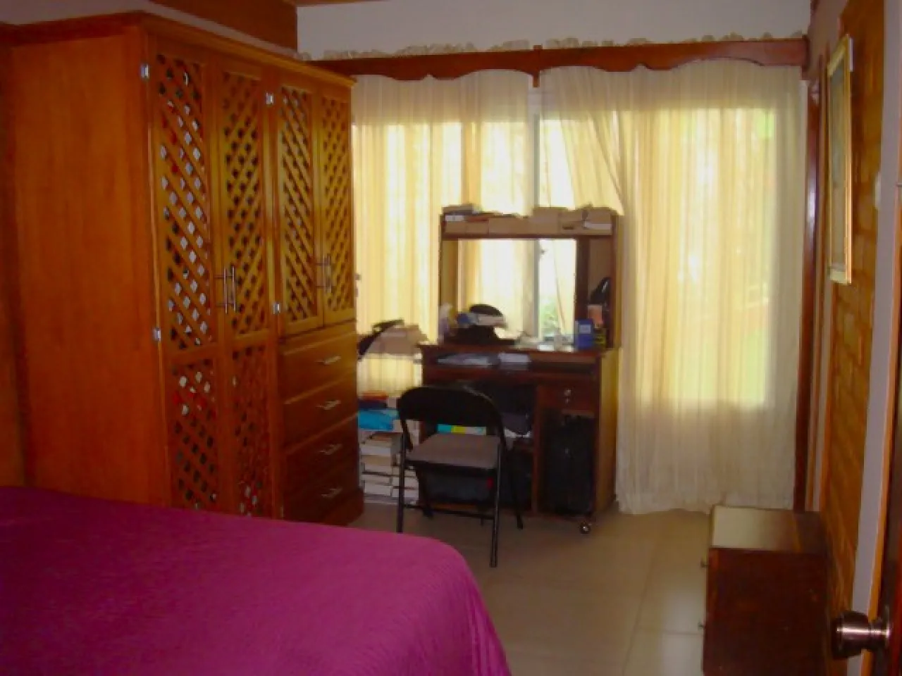 Habitación cuenta con cama matrimonial, clóset de madera, escritorio y silla de color negro, ventanas con cortinas blancas.