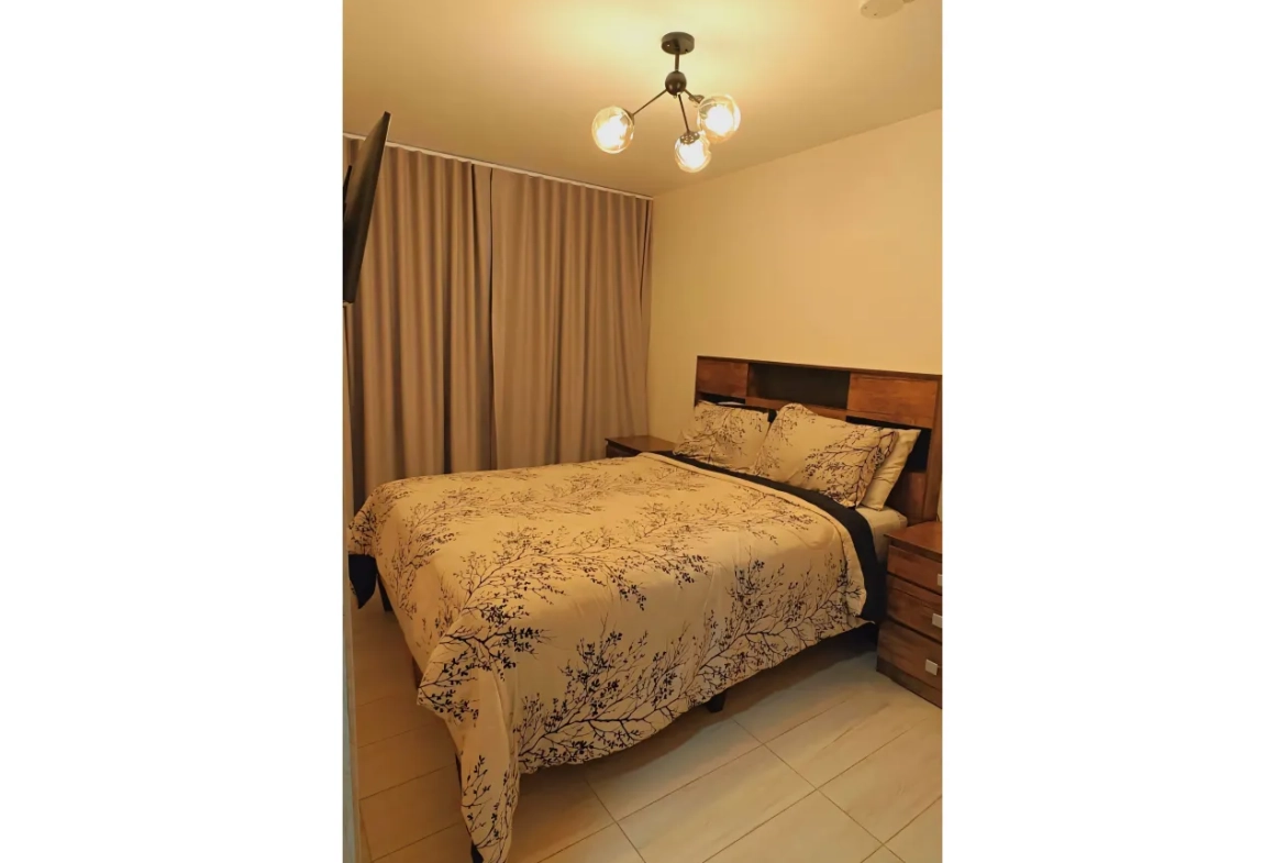 La habitación cuenta con una cama matrimonial con respaldo, además de una habitación con televisor y dos mesas de noche.