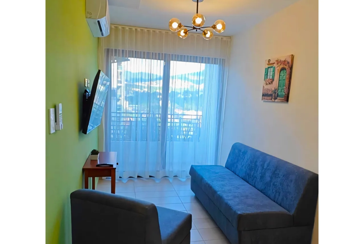 Sala de estar cuenta con paredes de color blanco combinadas con marillo, ademas la sala incluye dos sof+as, un televisor, una mesa de madera, aire acondicionado y acceso al área de balcón.