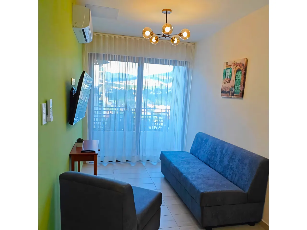 Sala de estar cuenta con paredes de color blanco combinadas con marillo, ademas la sala incluye dos sof+as, un televisor, una mesa de madera, aire acondicionado y acceso al área de balcón.