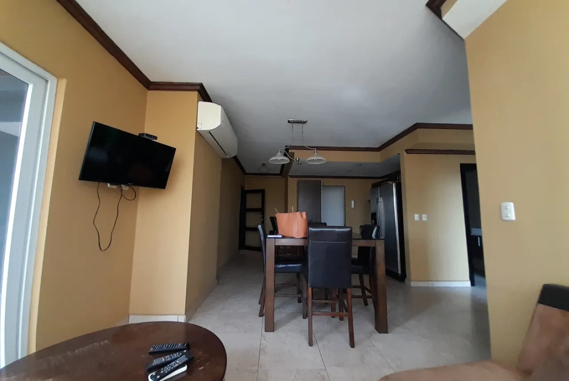 La sala de estar cuenta con sofá y una mesa de madera, de fondo podemos ver una mesa de madera con 4 sillas a juego, televisor colgado en la pared, aire acondicionado. Las paredes son de color amarillo oscuro.