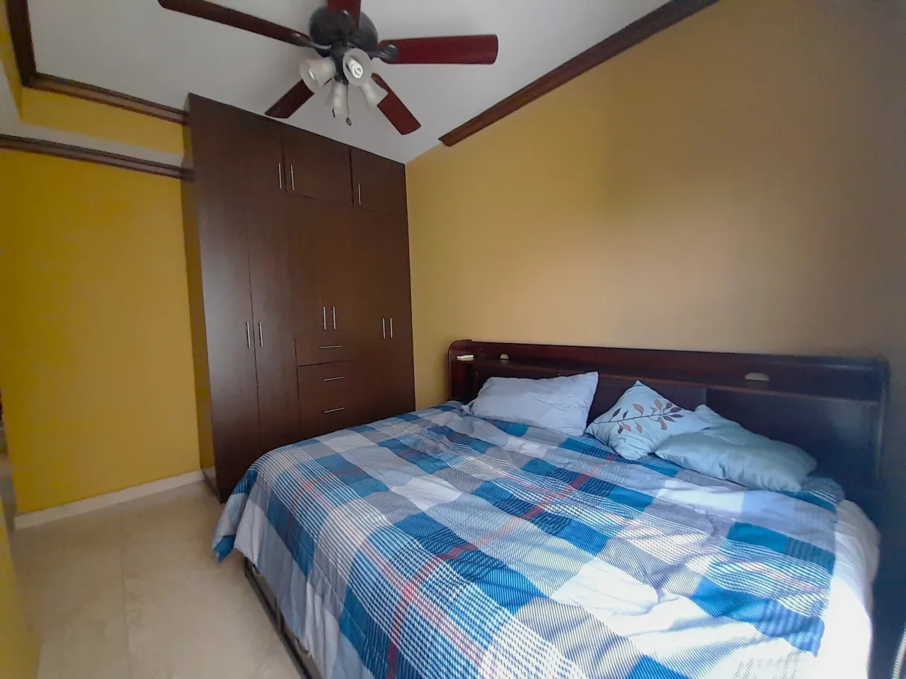 La habitación secundaria cuenta con una amplia cama matrimonial, clóset ventilador colgado en el techo, paredes de color amarillo y suelo de porcelanato.