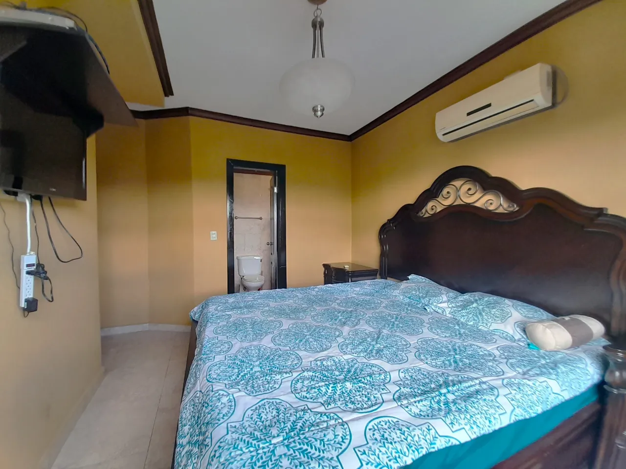Una de las habitaciones cuenta con una cama matrimonial, aire acondicionado, baño privado, paredes de color amarillo, televisor colgado en la pared.