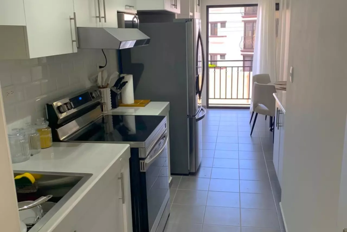 La cocina cuenta con muebles de color blanco, espacio para colocar electrodomesticos como estufa y refrigerador. De fondo podemos apreciar el acceso al balcón del apartamento.