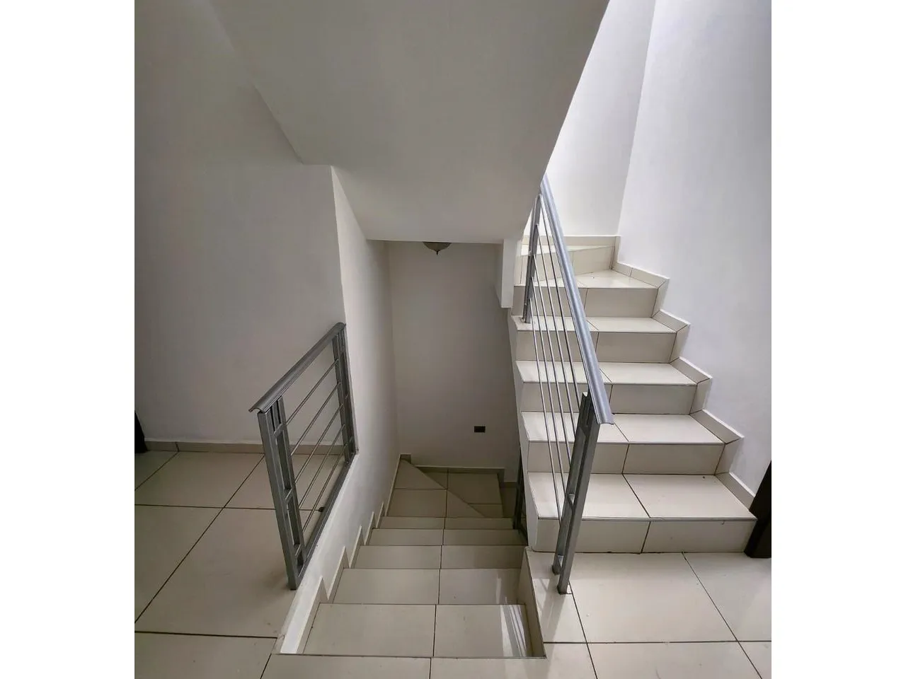 Escaleras que dan aceso al segundo nivel de la casa, cuenan con paredes de color blanco, suelo de porcelanato y los barandales son metalicos de color gris.
