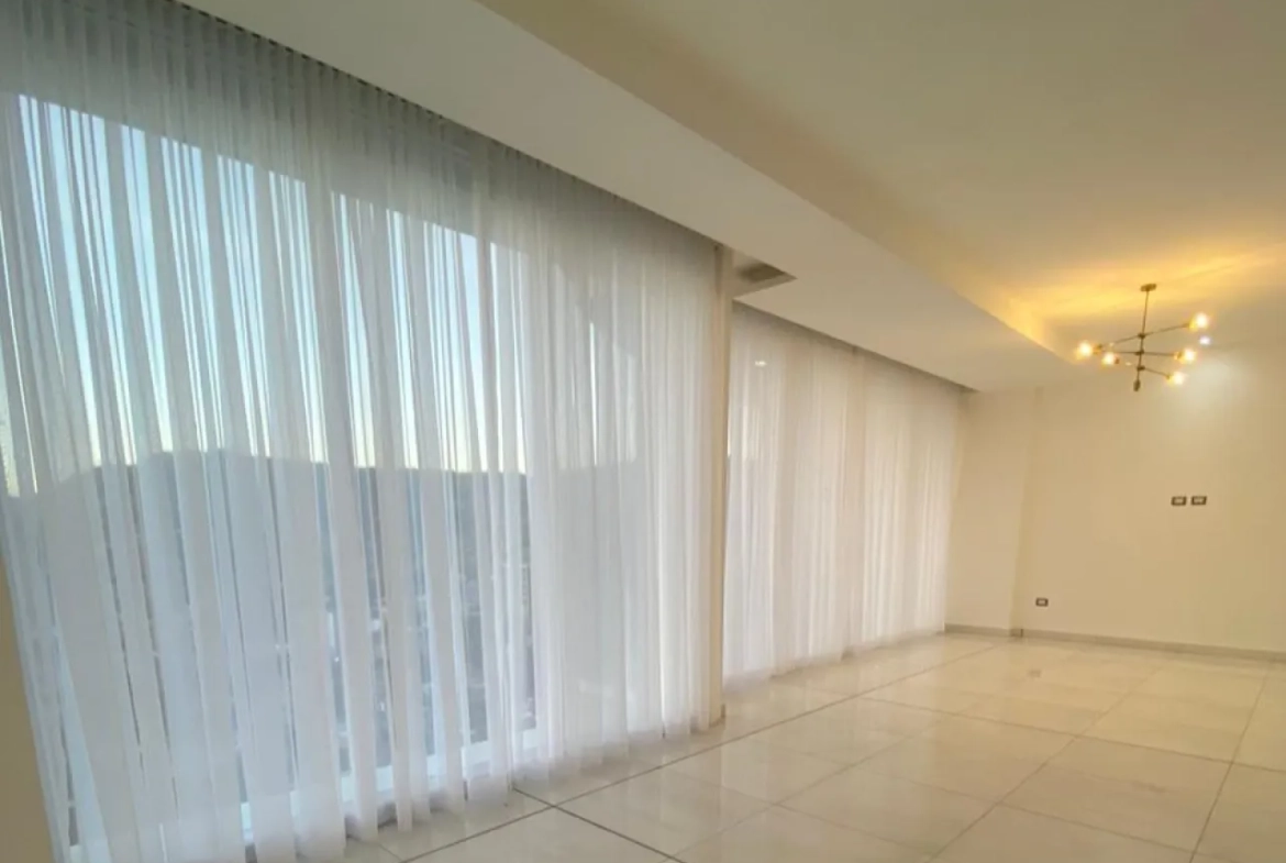 El apartamento cuenta con un área de sala y comedor amplios con cortinas automatizadas que brindan una hermosa vista de la ciudad. Las paredes son de color blanco y el suelo es de porcelanato.