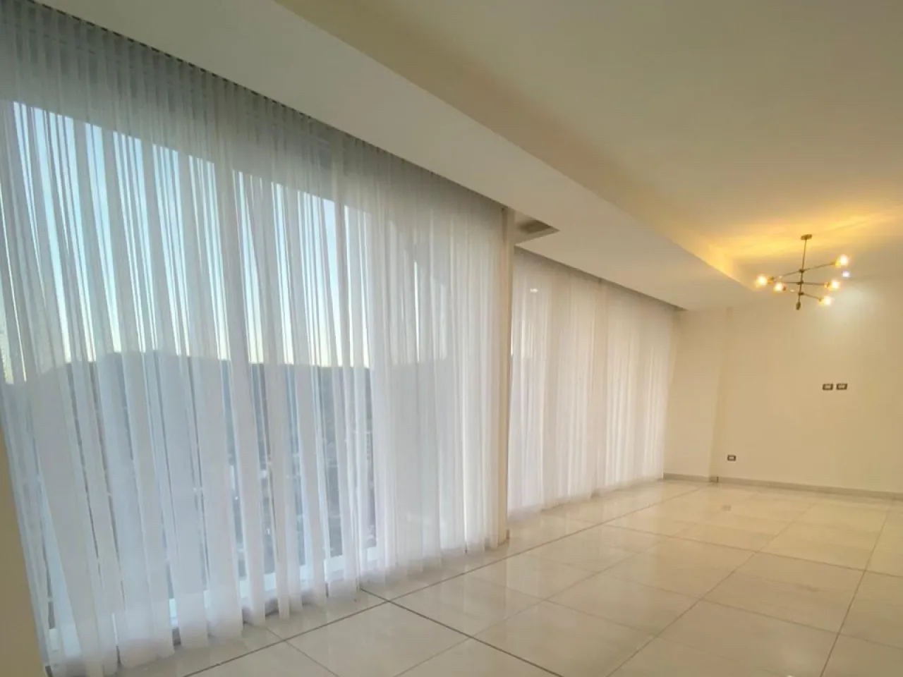 El apartamento cuenta con un área de sala y comedor amplios con cortinas automatizadas que brindan una hermosa vista de la ciudad. Las paredes son de color blanco y el suelo es de porcelanato.