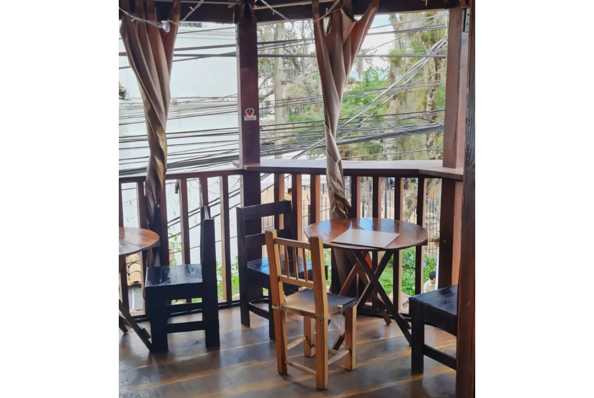Mesas redondas de madera, sillas de color negro a juego, balcón de madera con cortinas de color café.