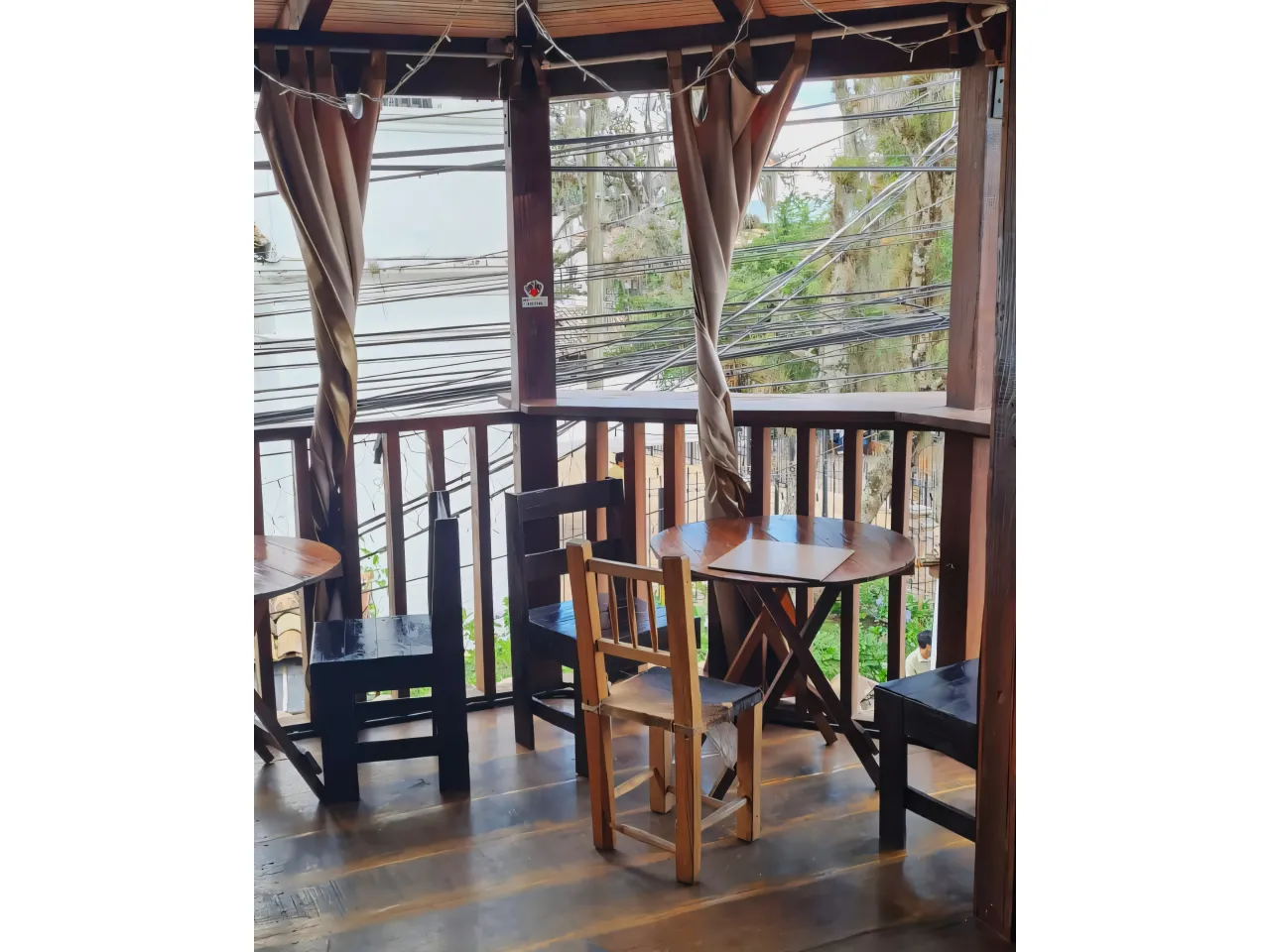 Mesas redondas de madera, sillas de color negro a juego, balcón de madera con cortinas de color café.