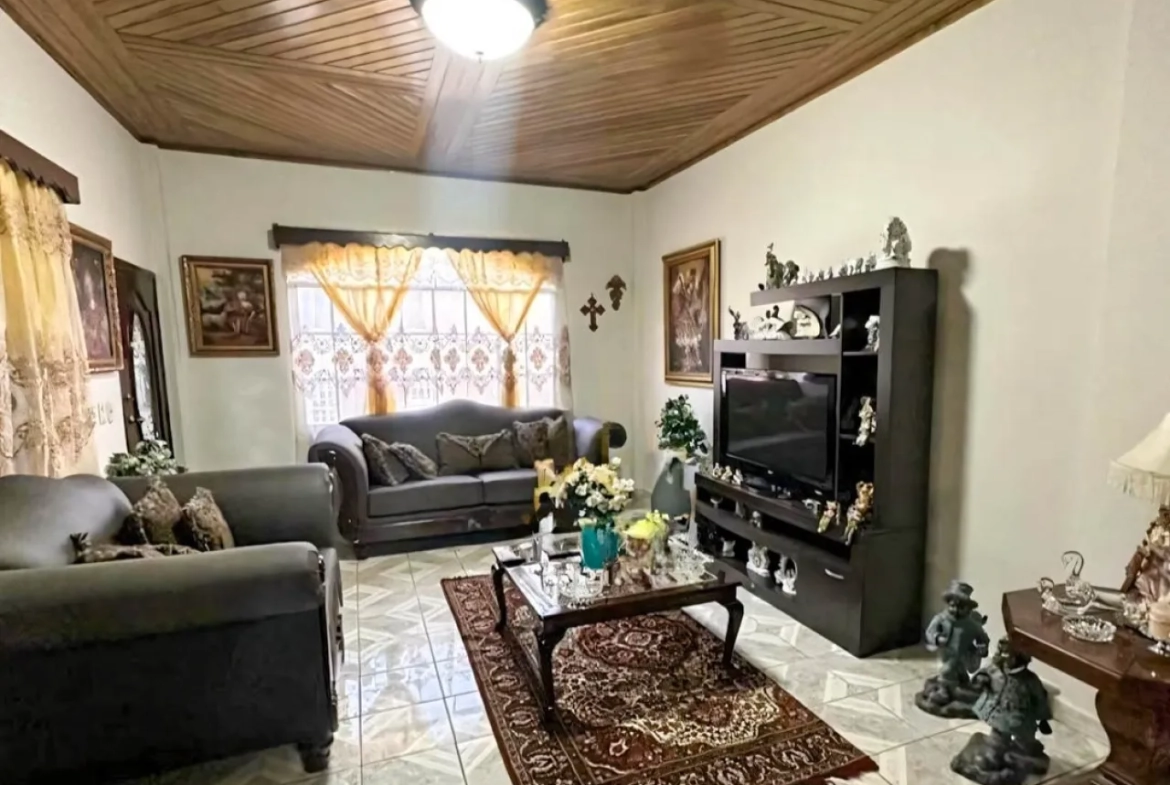 Sala de estar principal cuenta con sofas de color gris oscuro, con una alfombra que posse una mesa de vidrio encima, dos ventanas con cortinas amarillas y una división para colocar el televisor y diferentes muebles.