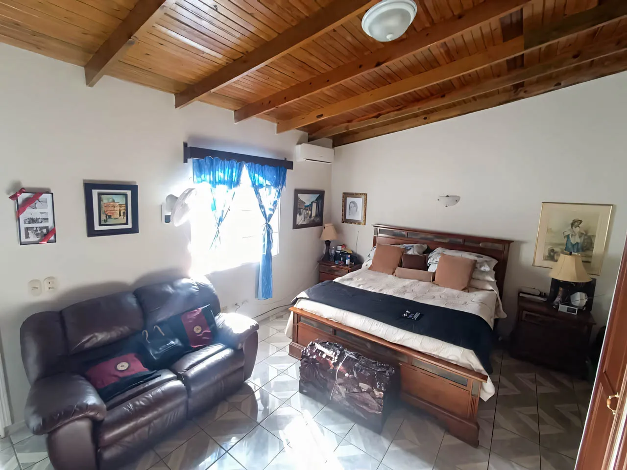 Una de las habitaciones cuenta con una amplia cama matrimonial, un sofá de cuerina, una ventana con cortinas de color azul que permiten una hermosa iluminación al interior.