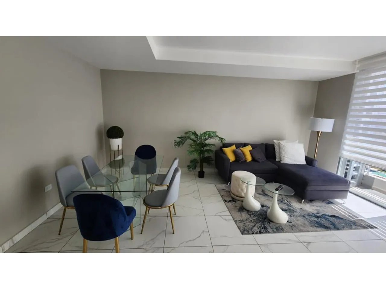 Espacio cuenta con sofá en forma largo de color gris oscuro, lámpara de color blanco con persianas en las ventanas, comedor rectangular de vidrio con 6 sillas