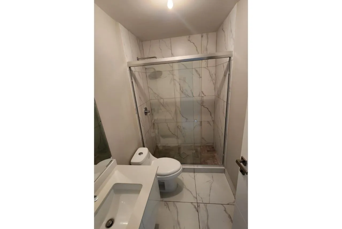 Uno de los baños cuenta con ducha con puertas corredizas de vidrio, retrete de color blanco y lavamanos con espejo amplio