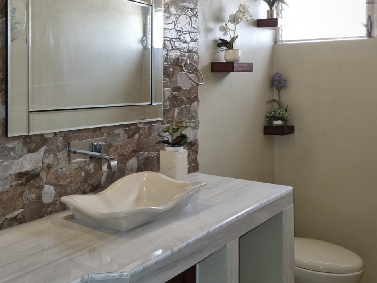 Baño moderno que cuenta con pared enchapada que lo acompaña un espejo elegante, lavamanos de ceramica y plantas para armonizar el espacio.