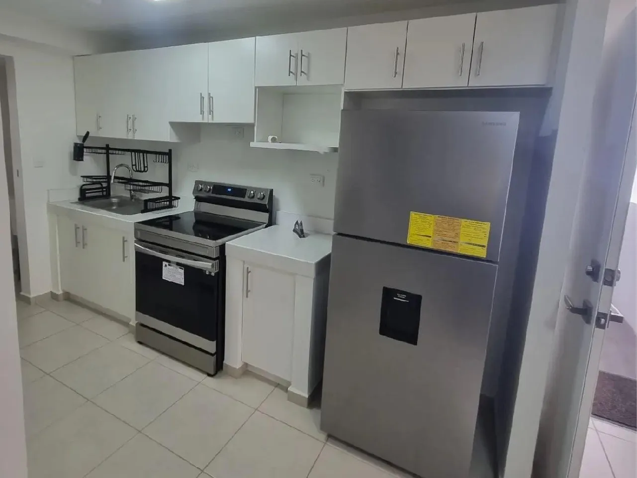 La cocina se encuentra totalmente equipada, cuenta con un refrigerador, estufa, escurridor de platos y muebles de color blanco.